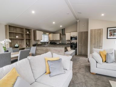 An open plan living room and kitchen with grey sofas with yellow and grey cushions a dining table with grey chairs and wooden kitchen cabinets at Parkers Place in Carnforth