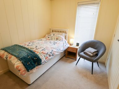 A small bedroom with a single bed a nightstand with a lamp a gray chair and a window with blinds at Tanqueray Lodge in Kenwick near Louth