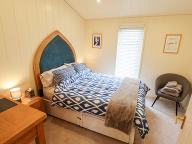 A bedroom with a bed with patterned bedding a side table with a lamp a gray chair with folded towels and framed pictures on the walls at Tanqueray Lodge Kenwick near Louth