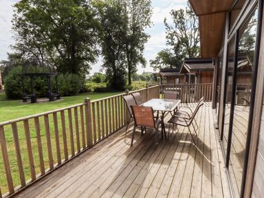 A wooden deck with a table and six chairs overlooking a green lawn with trees and wooden lodges at Tanqueray Lodge in Kenwick near Louth