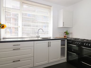 A kitchen with white cabinets a black oven a sink under a window and a bottle with glasses on the counter at Apartment at Morningside Hotel in Torquay