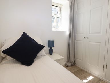 A bedroom with a single bed with white sheets and a dark pillow a small bedside table with a blue lamp a window with a model sailboat and closed white wardrobe doors at Apartment at Morningside Hotel in Torquay