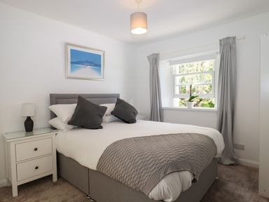 A bedroom with a bed with white and grey bedding a window with grey curtains a bedside table with a lamp and a framed picture on the wall at Apartment at Morningside Hotel in Torquay