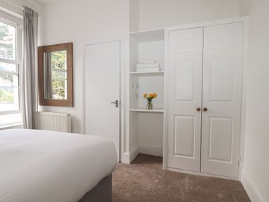 A bedroom with a bed near a window with curtains and a wooden framed mirror beside a white door and built-in shelves with towels and flowers at Apartment at Morningside Hotel in Torquay