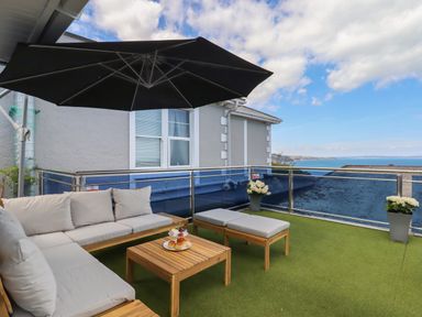 An outdoor seating area with a beige cushioned sofa a wooden table and a black umbrella overlooking the sea at Apartment at Morningside Hotel in Torquay