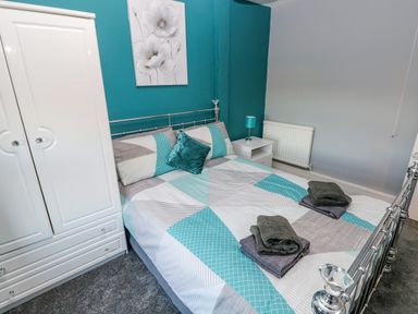 A bedroom with a patterned bedspread and folded towels on a bed with a metal frame a white wardrobe a nightstand with a lamp and a floral painting on a teal wall at Pant Teg Farm in Carmarthen
