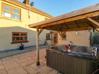 An outdoor patio area with a hot tub under a wooden pergola and a house with yellow walls in the background at Pant Teg Farm in Carmarthen