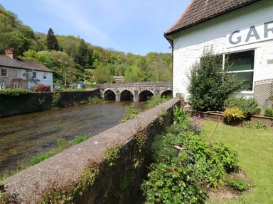 A river with a stone bridge and houses near a garden with plants and flowers at Barleside in Dulverton