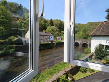 View through open windows overlooking a river with houses and a stone bridge surrounded by trees at Barleside in Dulverton