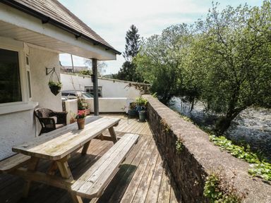 An outdoor wooden deck with a picnic table and potted plants next to a stone wall overlooking a river and trees at Barleside in Dulverton