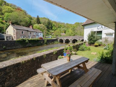 A wooden picnic table on a deck overlooking a river with a stone bridge and houses across the river at Barleside in Dulverton