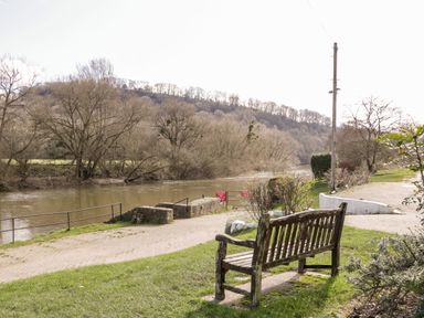 A wooden bench on grass facing a river with trees and a hill in the background at Steel Works Farm in Gorsley