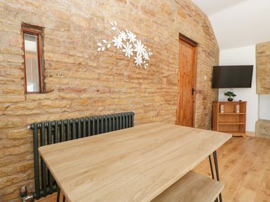 A dining area with a wooden table and benches against a stone wall with a radiator and mounted TV at Steel Works Farm in Gorsley