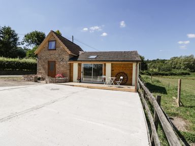 A small stone and wood house with a concrete driveway an outdoor table and chairs and a fenced grassy field at Steel Works Farm in Gorsley