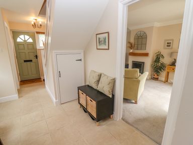 A hallway with a green front door a small bench with cushions and a cabinet with baskets a living room with a sofa fireplace and a plant visible through a doorway at The Cornerstone in Bourton-On-The-Water
