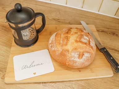 A loaf of bread with a bread knife a French press coffee maker and a welcome card on a wooden cutting board at The Cornerstone in Bourton-On-The-Water
