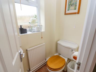 A small bathroom with a toilet a wooden seat a window with a windowsill radiator and a framed picture on the wall at The Cornerstone in Bourton-On-The-Water