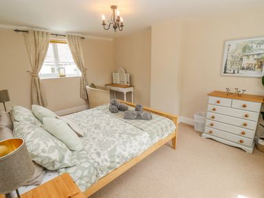 A bedroom with a wooden bed and patterned bedding a white dresser and a vanity table by a window at The Cornerstone in Bourton-On-The-Water