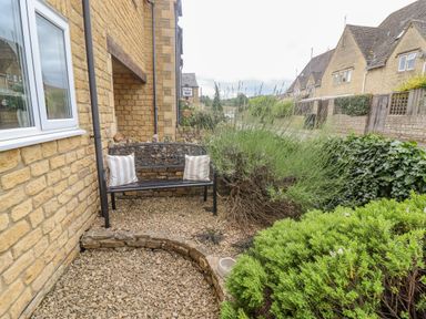 A small garden area with a metal bench with cushions and plants next to a stone building at The Cornerstone in Bourton-On-The-Water