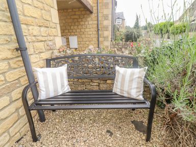 A metal bench with pillows and a welcome sign on a gravel surface outside a stone building at The Cornerstone in Bourton-On-The-Water