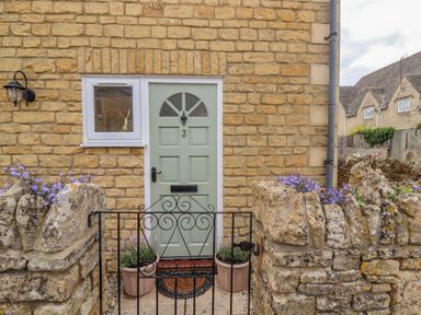 A front door with a small window and a gate surrounded by stone walls and flower pots at The Cornerstone in Bourton-On-The-Water