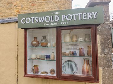 A display case with pottery items on shelves outside a building at The Cornerstone in Bourton-On-The-Water