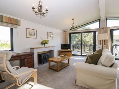 A living room with a white sofa green cushions a wooden coffee table a wooden desk and chair a fireplace and sliding glass doors at Manor Farm Lodge in Bowerchalke