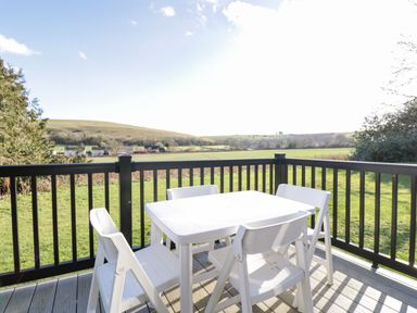 An outdoor deck with a white table and four chairs overlooking green fields and hills at Manor Farm Lodge in Bowerchalke