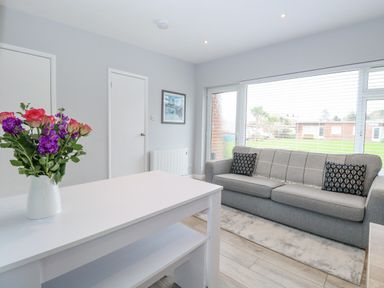 A room with a gray sofa with two pillows a white table with flowers and a large window at 12 Siesta Mar in Mundesley