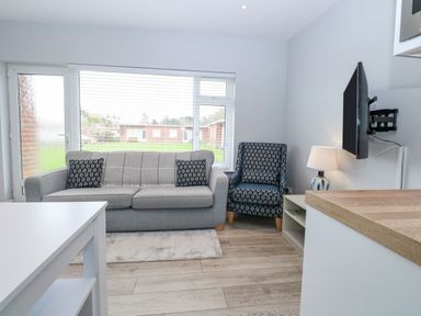 A living room with a grey sofa and patterned armchair a wall mounted TV and a lamp on a side table at 12 Siesta Mar in Mundesley