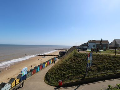 A beach with colorful beach huts and people on a promenade and grassy hill with houses in the background at 12 Siesta Mar in Mundesley