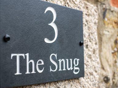 A close up of a black house number plaque with the number 3 and the words The Snug on a stone wall at The Snug in St Minver