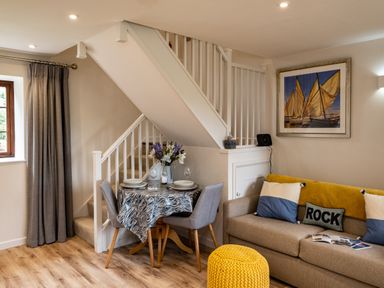 A dining table with two chairs next to a staircase and a sofa with cushions and a yellow knitted pouf in a living room at The Snug in St Minver