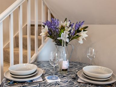 A dining table set for two with plates bowls wine glasses a bottle and flowers near stairs at The Snug in St Minver