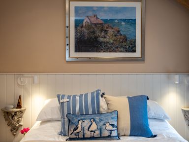 A bedroom with a bed with blue and beige pillows and a framed painting of a house by the sea above the bed at The Snug in St Minver