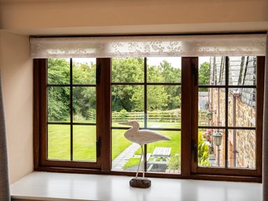 A wooden window with a white bird sculpture on the sill overlooking a garden with trees and a picnic table at The Snug in St Minver