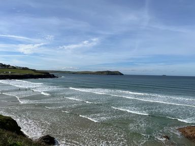 A beach with waves and surfers in the water with a headland and houses on the shore in the background at The Snug in St Minver