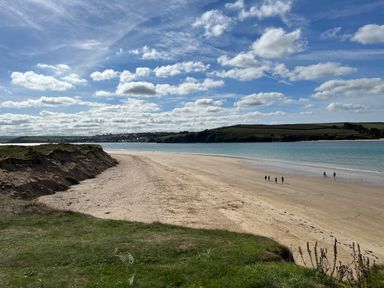 A sandy beach with a few people walking near the water and grassy dunes under a cloudy sky at The Snug in St Minver