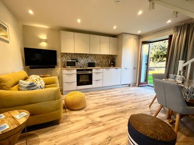 A kitchen and living area with a yellow sofa a wall-mounted TV white cabinets and an open door to a garden at The Snug in St Minver