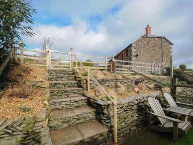 An outdoor stone staircase with wooden handrails leading up to a wooden fence and gate next to a stone building with wooden chairs at The Snug in St Minver