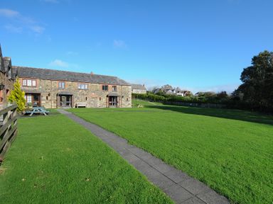 A stone house with wooden doors and windows facing a large lawn with a stone pathway and picnic tables at The Snug in St Minver