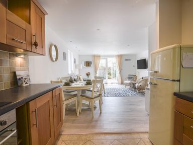 An open kitchen and dining area with wooden cabinets a round dining table and chairs leading into a living room with armchairs a sofa rug and a wall-mounted tv at 1 Buckfields in Lyme Regis