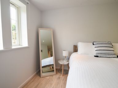 A bedroom with a wooden floor a window a standing mirror a bedside table with a lamp and a bed with white beddings and cushions at 1 Buckfields in Lyme Regis