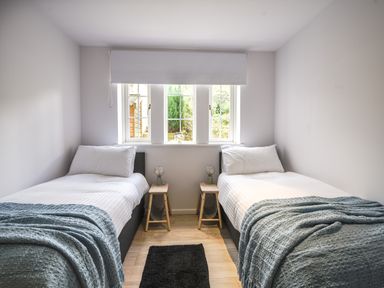 A bedroom with two single beds with white bedding and blue throws small wooden stools with lamps between the beds a window with a white roller blind at 1 Buckfields in Lyme Regis
