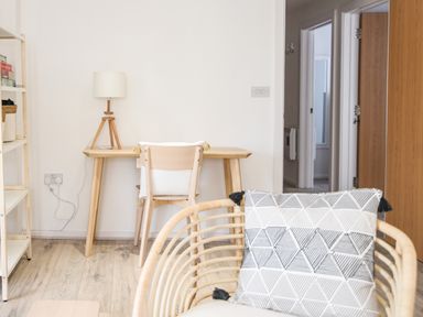 A room with a wooden desk and chair with a lamp a shelving unit and a rattan chair with a patterned cushion at 1 Buckfields in Lyme Regis