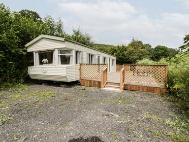 A mobile home with a wooden deck surrounded by trees and gravel at Grange Caravan in Llangollen