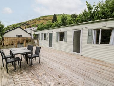 An outdoor wooden deck with a black table and four chairs next to a white caravan with green shutters at Grange Caravan in Llangollen