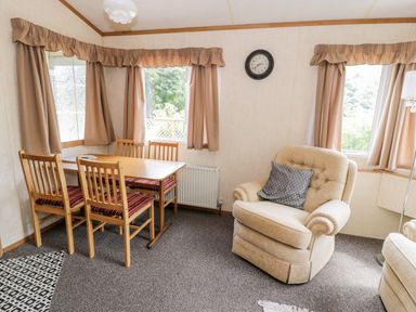 A dining area with wooden table and chairs next to an armchair with a cushion and windows with curtains at Grange Caravan in Llangollen