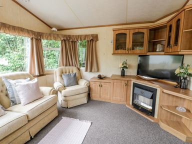 A living room with beige armchairs and a sofa with cushions a carpet in front and a wooden cabinet with a television and fireplace at Grange Caravan in Llangollen