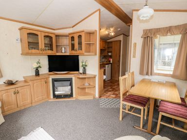 A living room with wooden cabinets a television a small dining table with chairs and a window with curtains at Grange Caravan in Llangollen